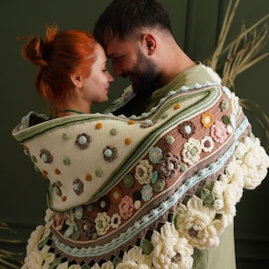 May include: A couple is standing close together, with their foreheads touching. They are both wearing a crocheted shawl with a floral pattern in shades of brown, green, and white. The shawl has a large, crocheted flower design around the edges.