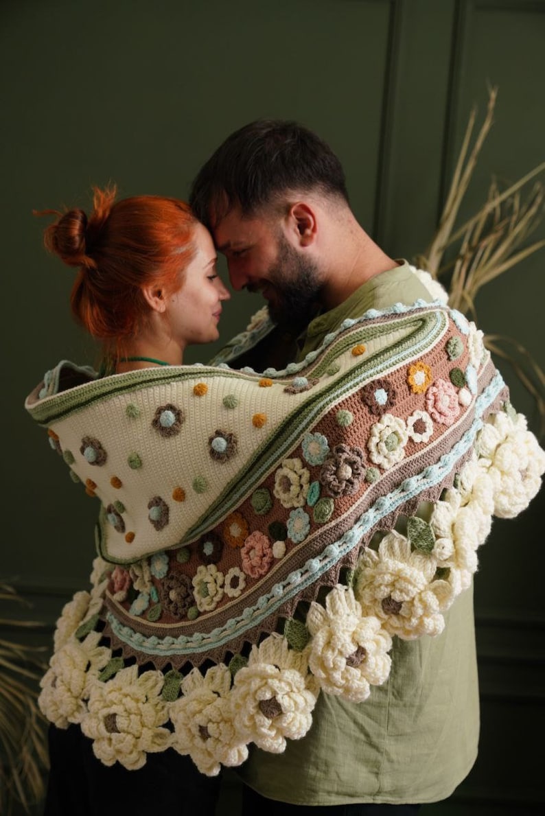 May include: A couple is standing close together, with their foreheads touching. They are both wearing a crocheted shawl with a floral pattern in shades of brown, green, and white. The shawl has a large, crocheted flower design around the edges.