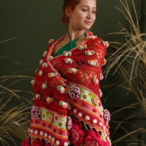 May include: A red crocheted shawl with colorful flowers and pom-poms. The shawl is draped over a person's shoulders and has a green lining.