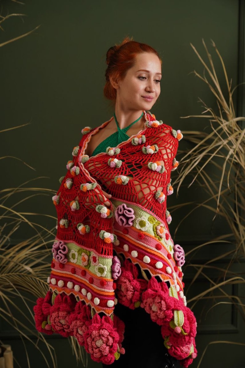 May include: A red crocheted shawl with colorful flowers and pom-poms. The shawl is draped over a person's shoulders and has a green lining.