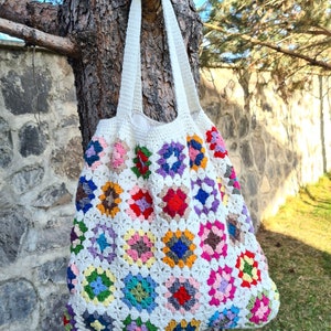 May include: A white crocheted tote bag with colorful granny squares. The bag is hanging on a tree trunk.