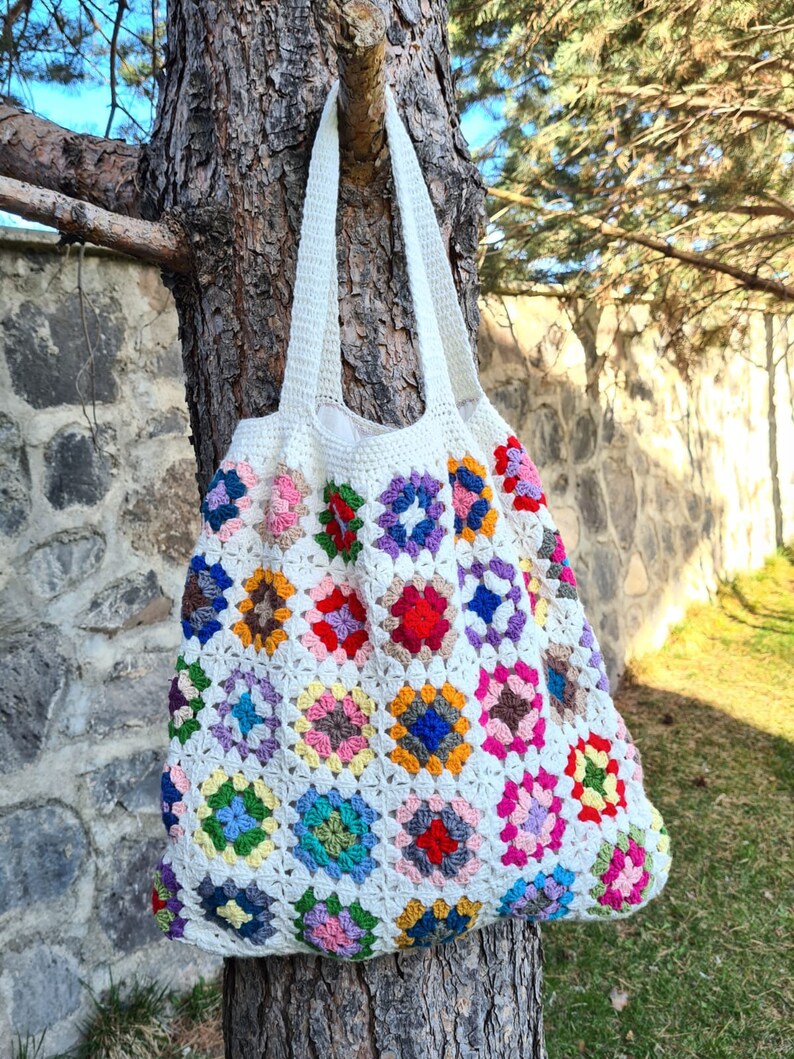 May include: A white crocheted tote bag with colorful granny squares. The bag is hanging on a tree trunk.