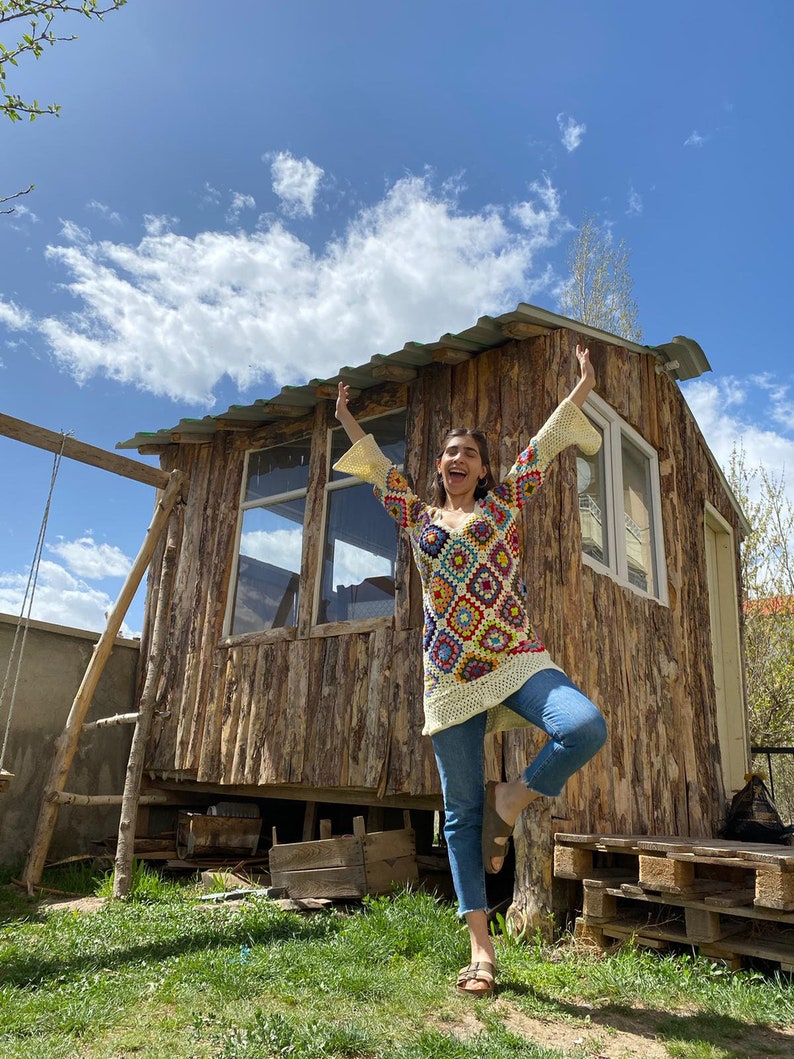 May include: A woman wearing a colorful crochet top with bell sleeves and blue jeans stands in front of a wooden cabin with a blue sky and white clouds in the background.