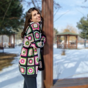 May include: A woman wearing a black crochet cardigan with colorful square patterns. The cardigan has a long, open front and is made with a chunky yarn.