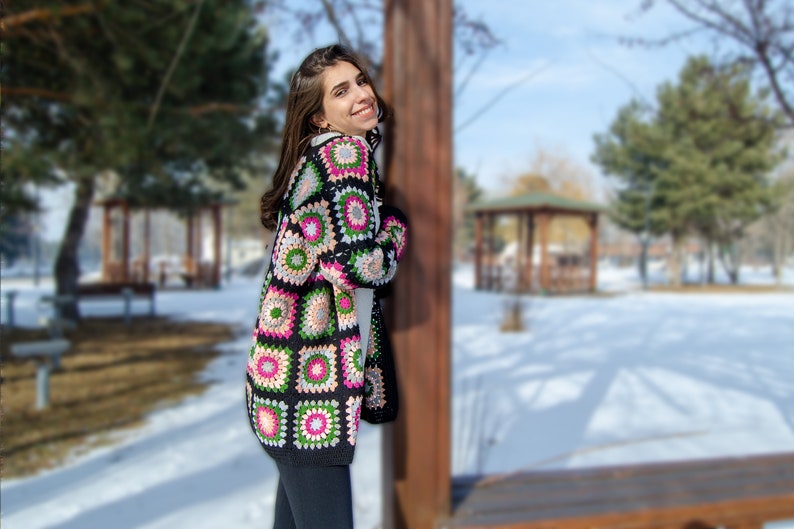 May include: A woman wearing a black crochet cardigan with colorful square patterns. The cardigan has a long, open front and is made with a chunky yarn.