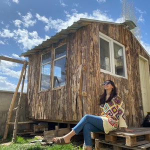 May include: A woman wearing sunglasses sits on a wooden pallet platform in front of a wooden cabin. She is wearing a crocheted top with a colorful geometric pattern and blue jeans.