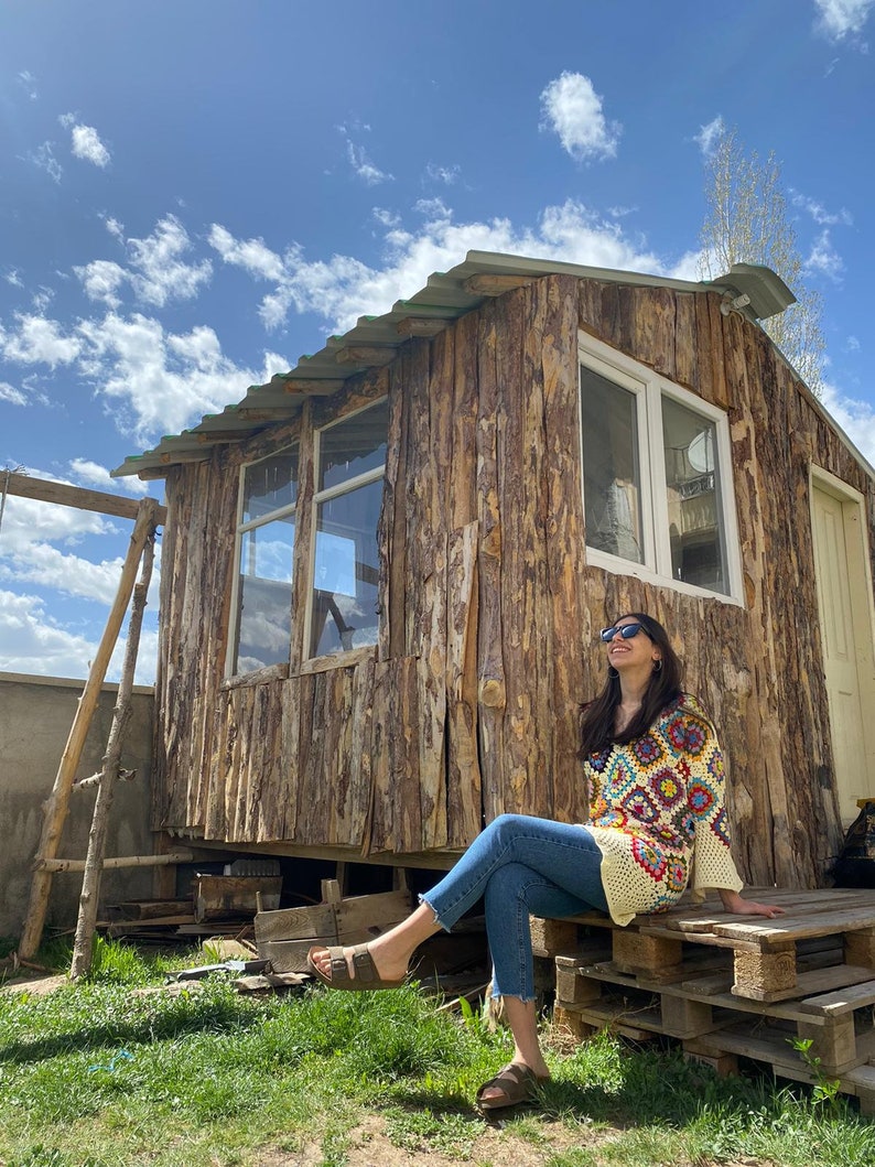 May include: A woman wearing sunglasses sits on a wooden pallet platform in front of a wooden cabin. She is wearing a crocheted top with a colorful geometric pattern and blue jeans.