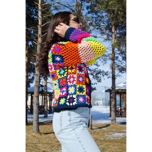 Puede incluir: Una chaqueta de ganchillo colorida con un dise&ntilde;o de patchwork que presenta cuadrados de varios colores, incluidos rosa, naranja, amarillo, azul, verde y morado. La chaqueta la lleva puesta una persona de pie en un entorno exterior nevado.