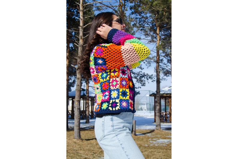 Puede incluir: Una chaqueta de ganchillo colorida con un dise&ntilde;o de patchwork que presenta cuadrados de varios colores, incluidos rosa, naranja, amarillo, azul, verde y morado. La chaqueta la lleva puesta una persona de pie en un entorno exterior nevado.
