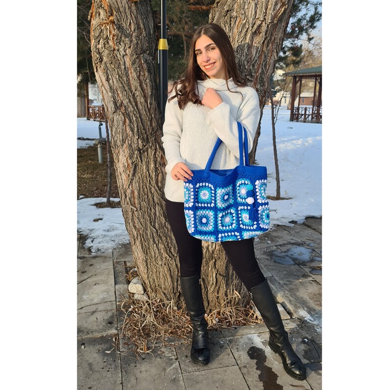 May include: A woman is standing in front of a tree holding a blue and white crocheted tote bag with pom poms. The bag has a square pattern.