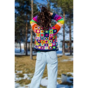 Puede incluir: Una mujer lleva una chaqueta de ganchillo colorida con un fondo azul y una variedad de colores brillantes. La chaqueta est&aacute; hecha de cuadrados con diferentes patrones. La mujer lleva pantalones vaqueros azules y est&aacute; de pie en un parque.