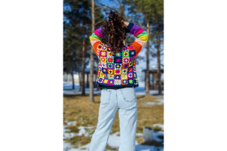 Puede incluir: Una mujer lleva una chaqueta de ganchillo colorida con un fondo azul y una variedad de colores brillantes. La chaqueta est&aacute; hecha de cuadrados con diferentes patrones. La mujer lleva pantalones vaqueros azules y est&aacute; de pie en un parque.