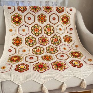 May include: A crocheted throw blanket with a geometric pattern of hexagons in shades of white, brown, orange, red, and green. The blanket has a fringe border and is draped over a white couch.