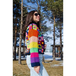 Puede incluir: Una mujer lleva una chaqueta de punto de ganchillo de colores con un patr&oacute;n de rayas arco&iacute;ris. La chaqueta est&aacute; hecha de cuadrados de diferentes colores y tiene un fondo negro. La mujer lleva pantalones vaqueros azules y gafas de sol.