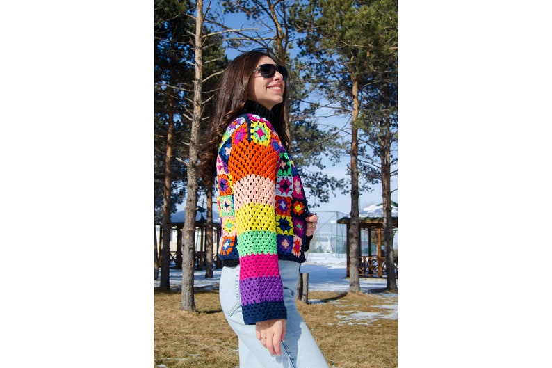 Puede incluir: Una mujer lleva una chaqueta de punto de ganchillo de colores con un patr&oacute;n de rayas arco&iacute;ris. La chaqueta est&aacute; hecha de cuadrados de diferentes colores y tiene un fondo negro. La mujer lleva pantalones vaqueros azules y gafas de sol.