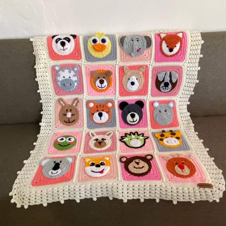 Puede incluir: Una manta de beb&eacute; de crochet blanco con un borde rosa y gris. La manta presenta una variedad de aplicaciones de animales, incluyendo un panda, un pato, un elefante, un zorro, una cebra, un le&oacute;n, un canguro, un perro, un conejo, un tigre, un oso, una jirafa, una rana, un koala, un zorro y un ciervo.