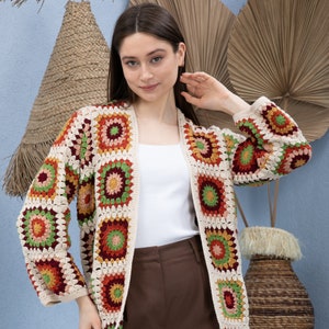 Pu&ograve; includere: Un cardigan all'uncinetto con sfondo crema e motivi geometrici colorati. Il cardigan ha un'apertura anteriore lunga e maniche lunghe.