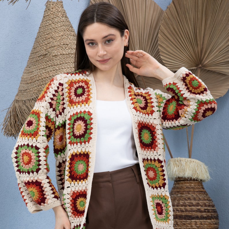 Pu&ograve; includere: Un cardigan all'uncinetto con sfondo crema e motivi geometrici colorati. Il cardigan ha un'apertura anteriore lunga e maniche lunghe.