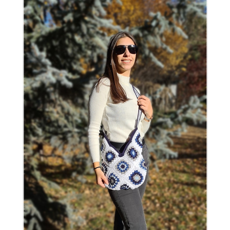 May include: A white crocheted shoulder bag with blue and gray geometric patterns. The bag has a long strap and is worn by a person wearing a white sweater and jeans.