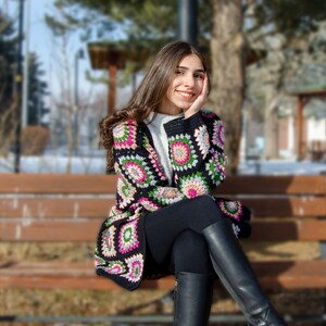 May include: A woman wearing a colorful crocheted cardigan with black pants and boots sits on a wooden bench in a snowy outdoor setting.