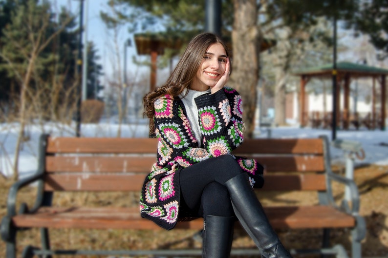 May include: A woman wearing a colorful crocheted cardigan with black pants and boots sits on a wooden bench in a snowy outdoor setting.
