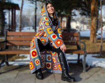 May include: A long, gray crochet cardigan with a colorful geometric pattern. The cardigan has a hood and is worn by a person sitting on a wooden bench.
