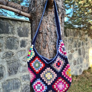 May include: A crocheted tote bag with a blue background and colorful granny squares. The bag has a long strap and is hanging on a tree trunk.