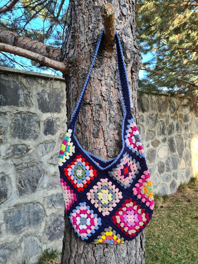 May include: A crocheted tote bag with a blue background and colorful granny squares. The bag has a long strap and is hanging on a tree trunk.