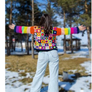 Puede incluir: Una mujer lleva una chaqueta de ganchillo de colores con un patr&oacute;n de rayas arco&iacute;ris. La chaqueta est&aacute; hecha de cuadrados de la abuela y tiene un fondo azul. La mujer lleva pantalones vaqueros azules claros y est&aacute; de pie en un bosque nevado.