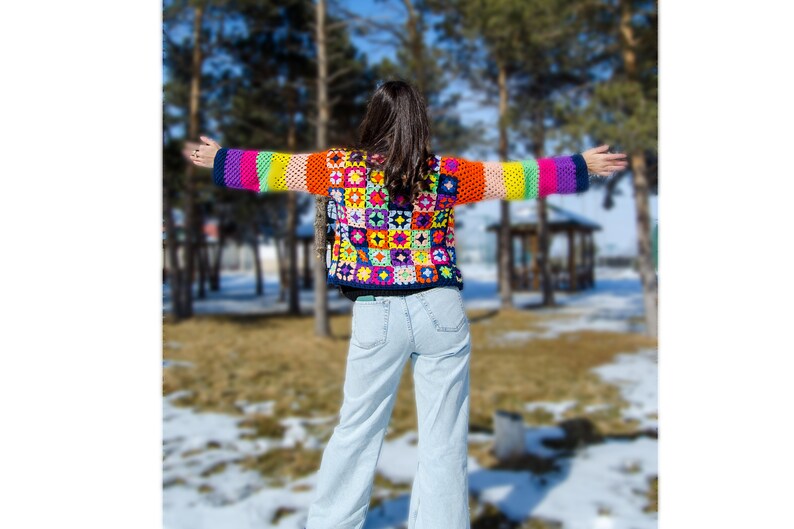 Puede incluir: Una mujer lleva una chaqueta de ganchillo de colores con un patr&oacute;n de rayas arco&iacute;ris. La chaqueta est&aacute; hecha de cuadrados de la abuela y tiene un fondo azul. La mujer lleva pantalones vaqueros azules claros y est&aacute; de pie en un bosque nevado.