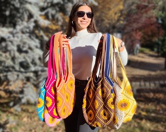 May include: A collection of crocheted tote bags with sunflower designs in various colors, including pink, blue, yellow, orange, brown, and white. The bags have a square shape with a diamond pattern and are held by a woman wearing sunglasses.