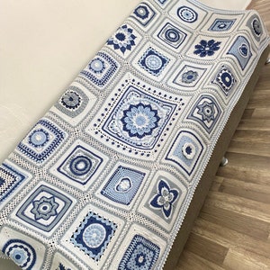 May include: A white crocheted blanket with blue and grey squares featuring various floral and geometric patterns.