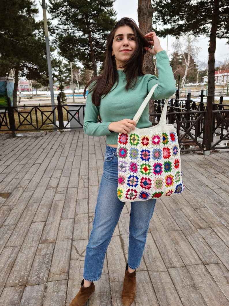 May include: A woman is holding a white crochet tote bag with colorful squares. The bag has a long strap and is made of thick yarn. The woman is wearing a green sweater and blue jeans.