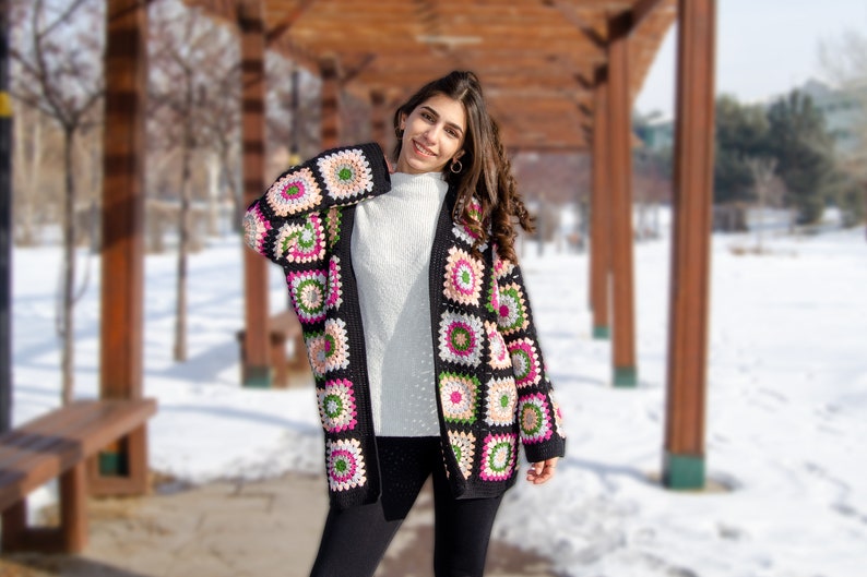 May include: A woman wearing a black crocheted cardigan with colorful squares. The cardigan has a long, loose fit and is made with a chunky yarn. The woman is standing in a snowy park setting.