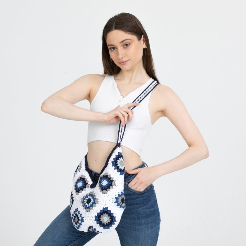 May include: A white crocheted tote bag with blue and gray floral designs. The bag has a long strap and is worn over the shoulder.