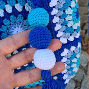 May include: A blue crocheted bag with a white, blue, and turquoise pattern. The bag has a blue tassel and three crocheted pom-poms in blue, white, and turquoise.