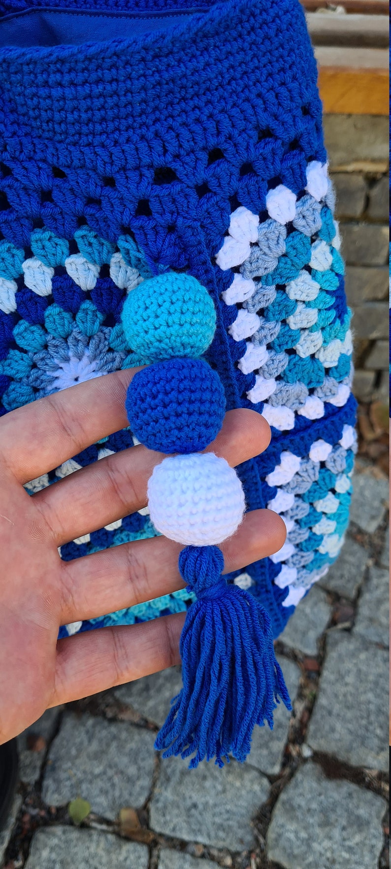 May include: A blue crocheted bag with a white, blue, and turquoise pattern. The bag has a blue tassel and three crocheted pom-poms in blue, white, and turquoise.