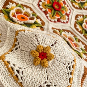 May include: A close-up of a crocheted blanket with a floral design. The blanket is made with white, brown, yellow, orange, and red yarn. The center of the blanket features a large flower with a red center and yellow petals.