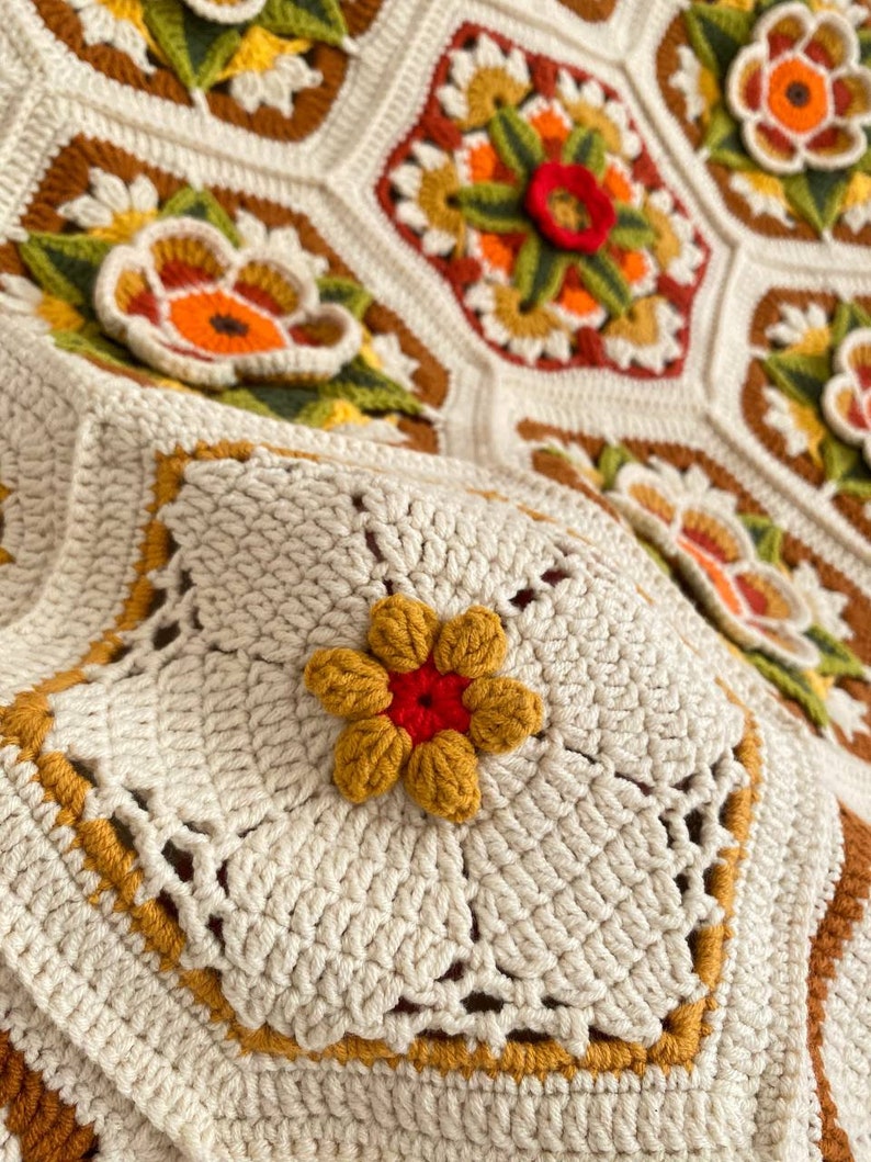 May include: A close-up of a crocheted blanket with a floral design. The blanket is made with white, brown, yellow, orange, and red yarn. The center of the blanket features a large flower with a red center and yellow petals.