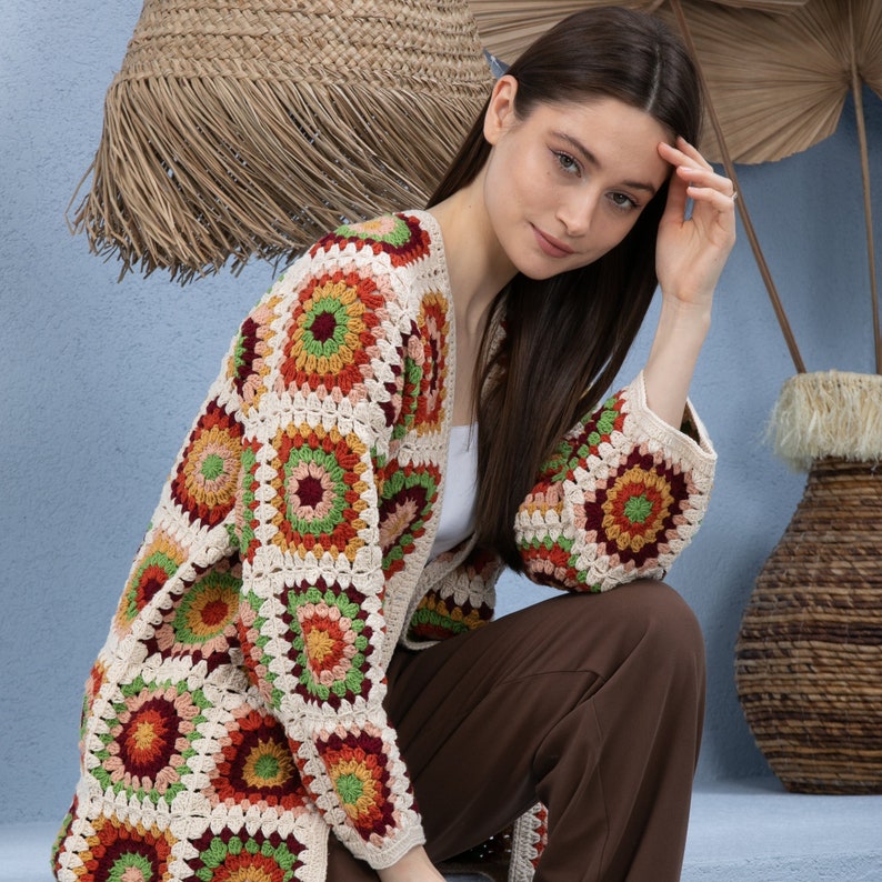 Pu&ograve; includere: Una donna indossa un cardigan all'uncinetto con un motivo geometrico colorato. Il cardigan &egrave; aperto sul davanti e ha maniche lunghe. La donna indossa pantaloni marroni e ha lunghi capelli castani.