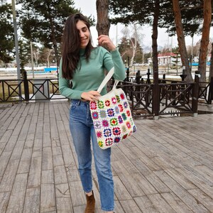 May include: A woman wearing a light green turtleneck sweater and blue jeans is holding a white crocheted tote bag with colorful squares. The bag has a variety of colors including red, yellow, blue, green, and purple.