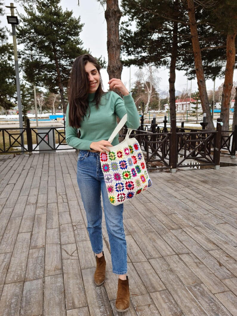 May include: A woman wearing a light green turtleneck sweater and blue jeans is holding a white crocheted tote bag with colorful squares. The bag has a variety of colors including red, yellow, blue, green, and purple.