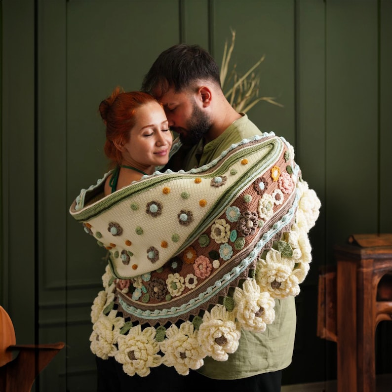 May include: A couple is wrapped in a large, crocheted shawl with a floral design. The shawl is cream, brown, and green with white flowers. The couple is standing in front of a green wall.