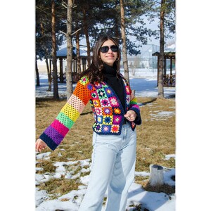 Puede incluir: Una chaqueta de crochet colorida con un patr&oacute;n de rayas arco&iacute;ris en las mangas. La chaqueta est&aacute; hecha de una variedad de colores, incluyendo rojo, naranja, amarillo, verde, azul, morado y rosa.