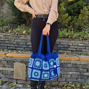 May include: A blue crocheted tote bag with white and light blue squares. The bag has a tassel and white pom-poms.