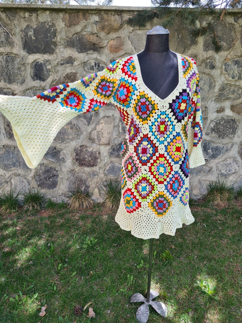 May include: A crocheted dress with a granny square pattern in various colors, including red, blue, green, yellow, and white. The dress has a V-neck and long sleeves with bell bottoms.