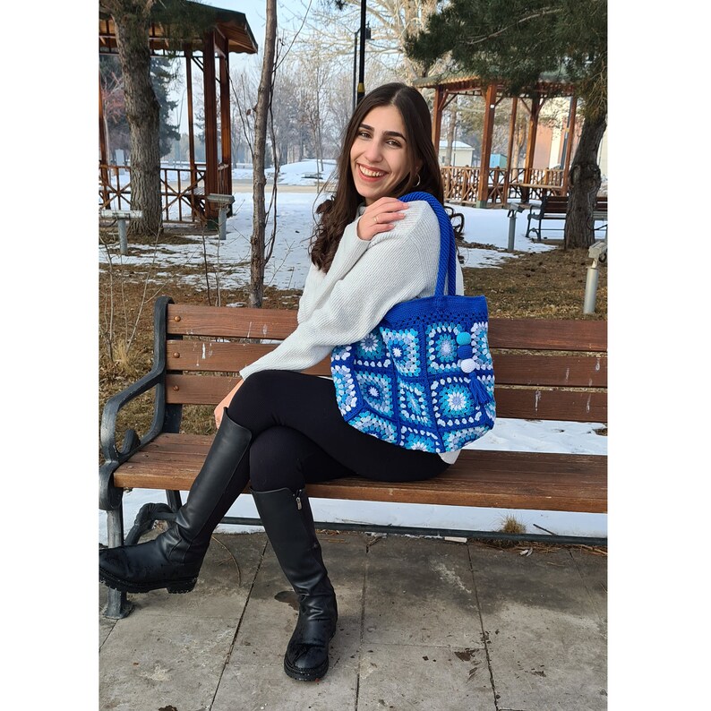 May include: A woman sits on a wooden bench wearing black trousers and boots, holding a blue and white crocheted tote bag with white pom-poms.