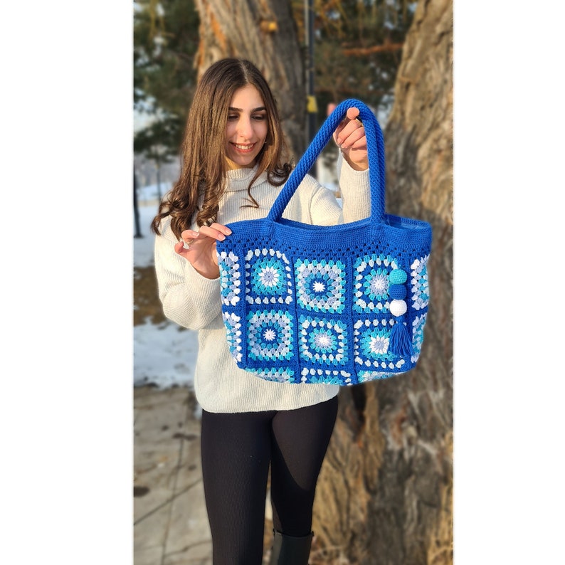 May include: A blue crocheted tote bag with white and light blue accents. The bag has a square shape and features a tassel.