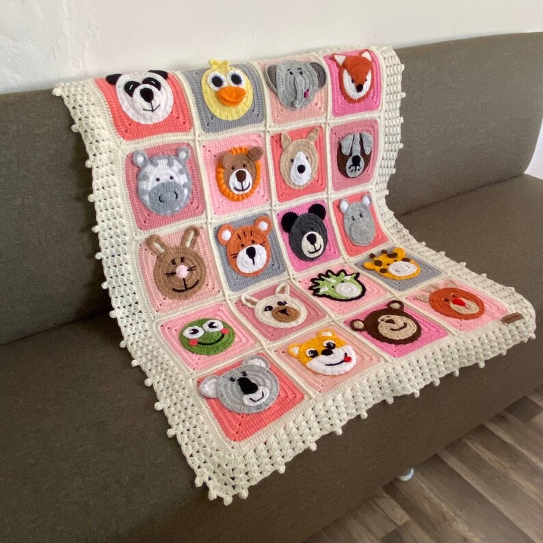 Puede incluir: Una manta de beb&eacute; de crochet con un borde blanco y un fondo rosa y gris. La manta presenta una variedad de caras de animales, incluyendo un panda, una cebra, un le&oacute;n, un tigre, un mono, una jirafa, una rana, un erizo, un koala y un zorro.