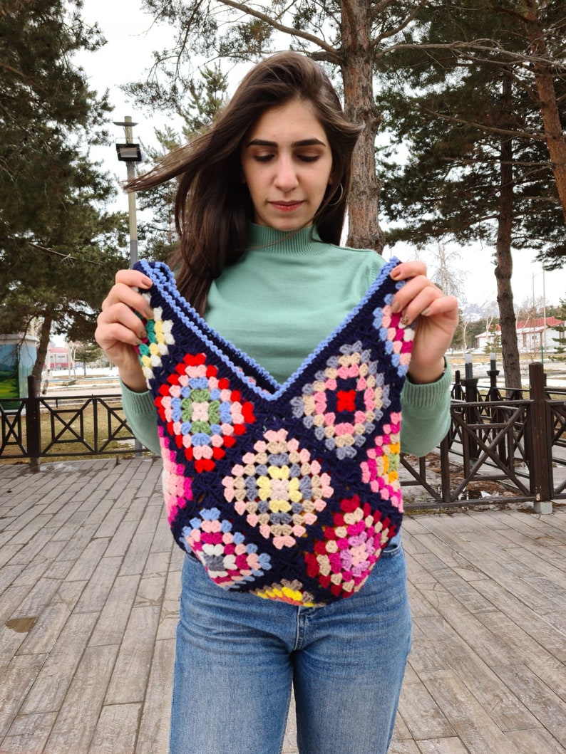 May include: A woman is holding a crocheted tote bag with a colorful granny square pattern. The bag is made with blue, pink, red, yellow, green, and white yarn.